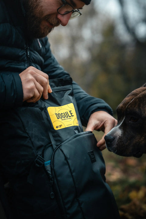 Muž vytahující psí pamlsky Dogule z jeho cestovního batohu vedle jeho psa